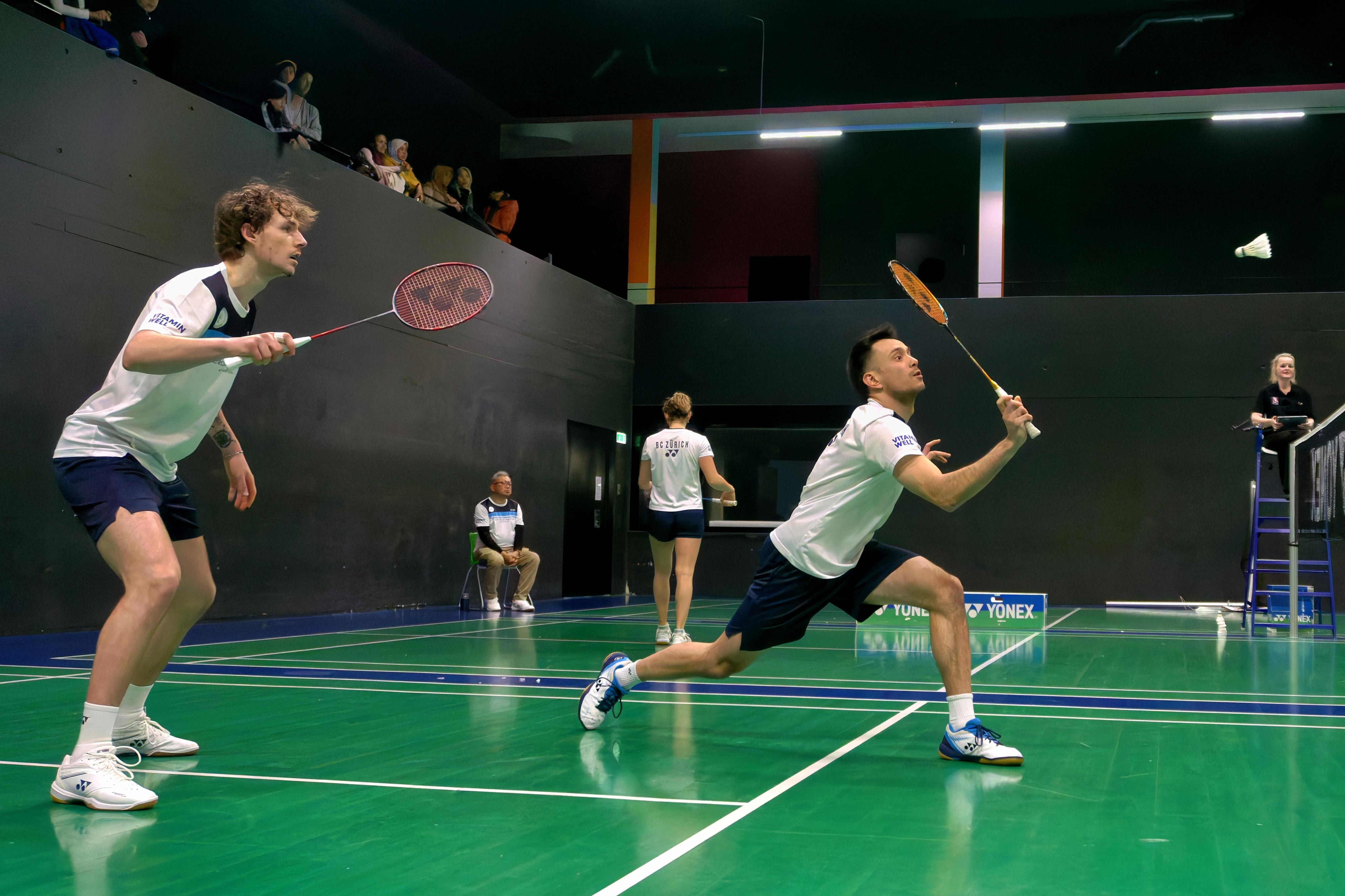 Zach Russ & Nicolas A. Müller spielen einen Punkt in einem NLA Doppel in einem Heimspiel in der Yonex Badminton Halle Letzigrund. Im Hintergrund spielt Lucie Amiguet ihr Einzel.